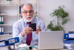 Starszy mężczyzna przed laptopem. W ręku trzyma telefon, patrzy na wyświetlacz