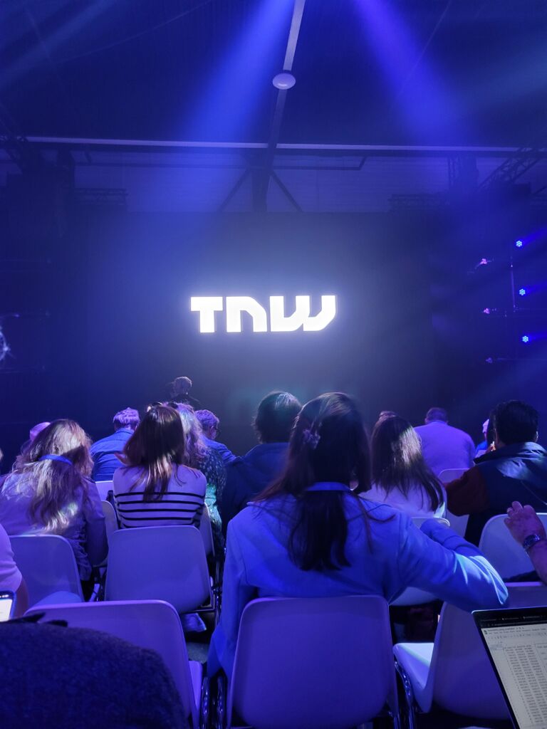 Ludzie siedzący na konferencji. Na ekranie wyświetla się TNW. Przyciemnione światło.