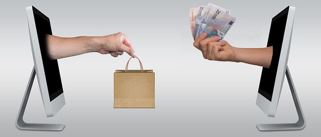 dwie ręce na przeciwko siebie wystające z monitorów tabletów. Jedna ręka trzyma plik pieniędzy, druga ręka trzyma papierową torbę z zakupami