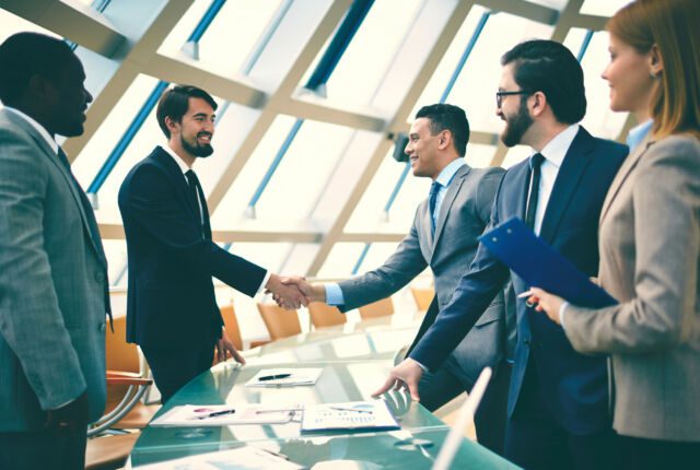 czterech mężczyzn i jednak kobieta w sali konferencyjnej. Dwóch mężczyzn ściska sobie ręce. Na stole wykresy i raporty.